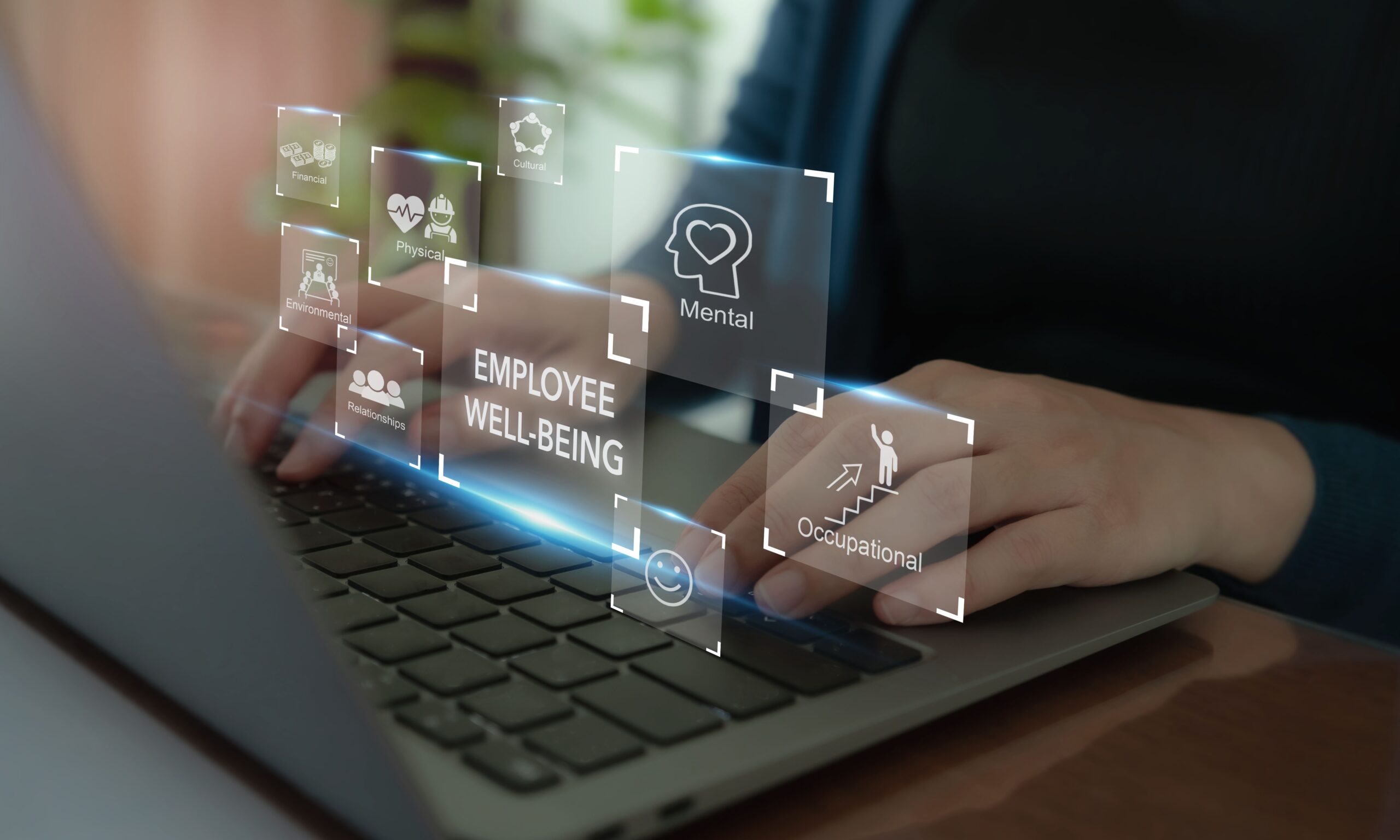 A person is sitting in front of a keyboard typing. There are text boxes floating with writing on them, containing symbols and words related to employee well-being, including mental, occupational, relationships, environmental, physical, and financial.